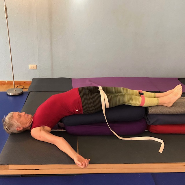 Yoga for immune system: Chris Lalor lying back over stacked bolsters in a supported bridge pose, with a belt around her thighs and her arms resting on the floor