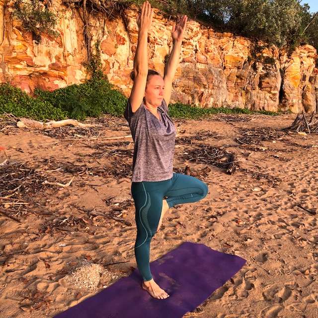 Kim in Tree Pose, Vrksasana, arms raised, standing on a beach