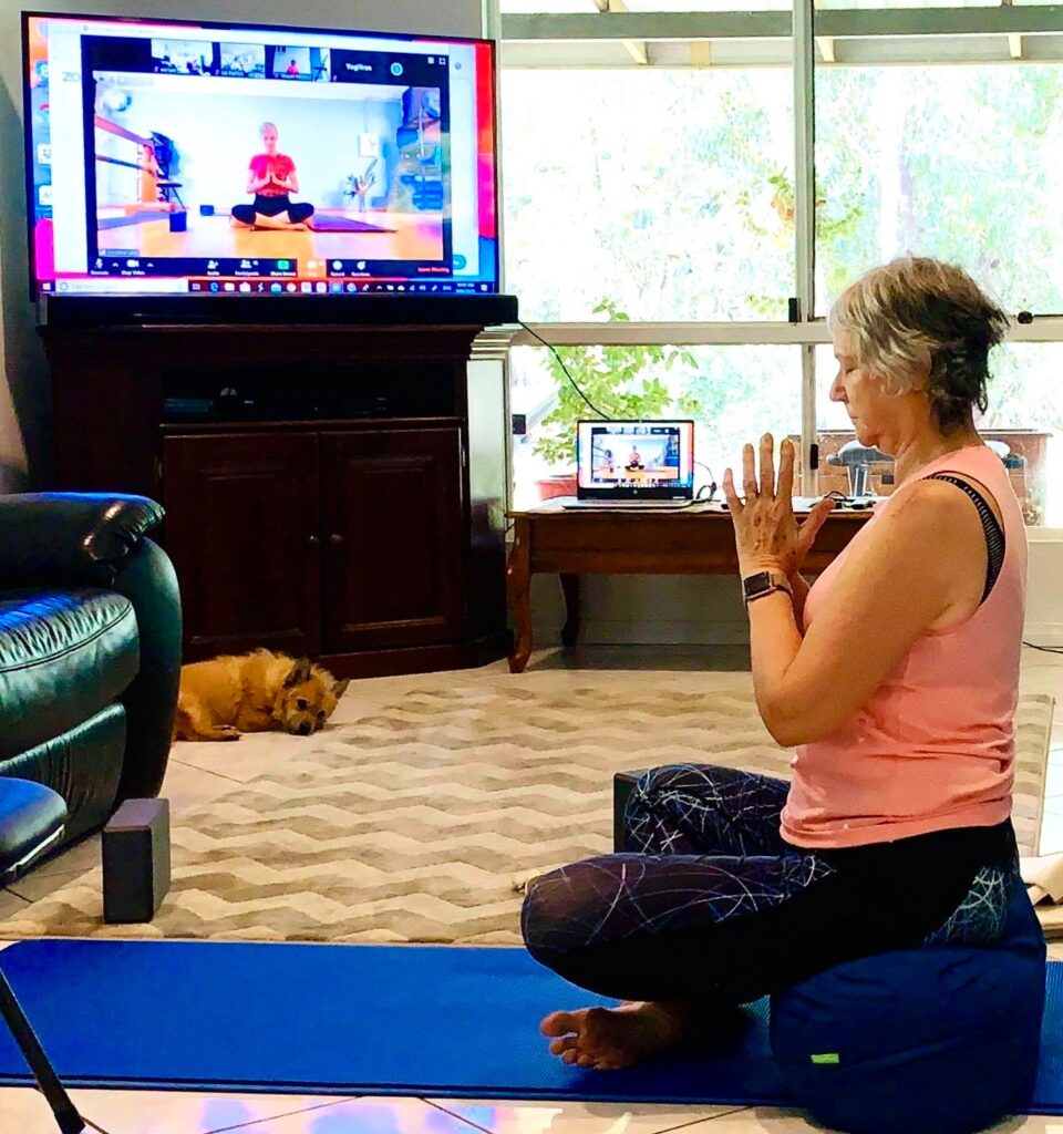 A Flametree student practising Iyengar yoga online at home, seated on a bolster in front of her TV, with her dog asleep nearby