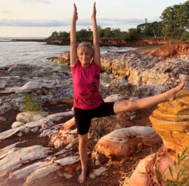 Chris Lalor in a standing balance pose on red rock at the water's edge near Darwin at sunset, arms raised overhead, one leg extended to the side.