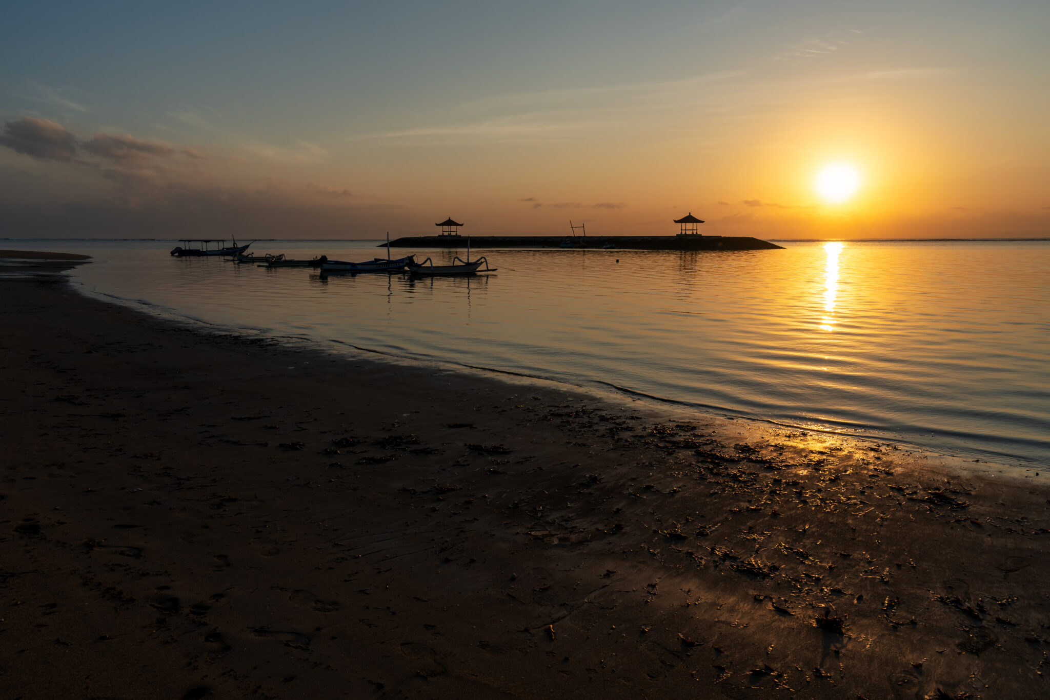 The beach at Sanur, Bali, with the sun low over calm water and outrigger boats moored offshore