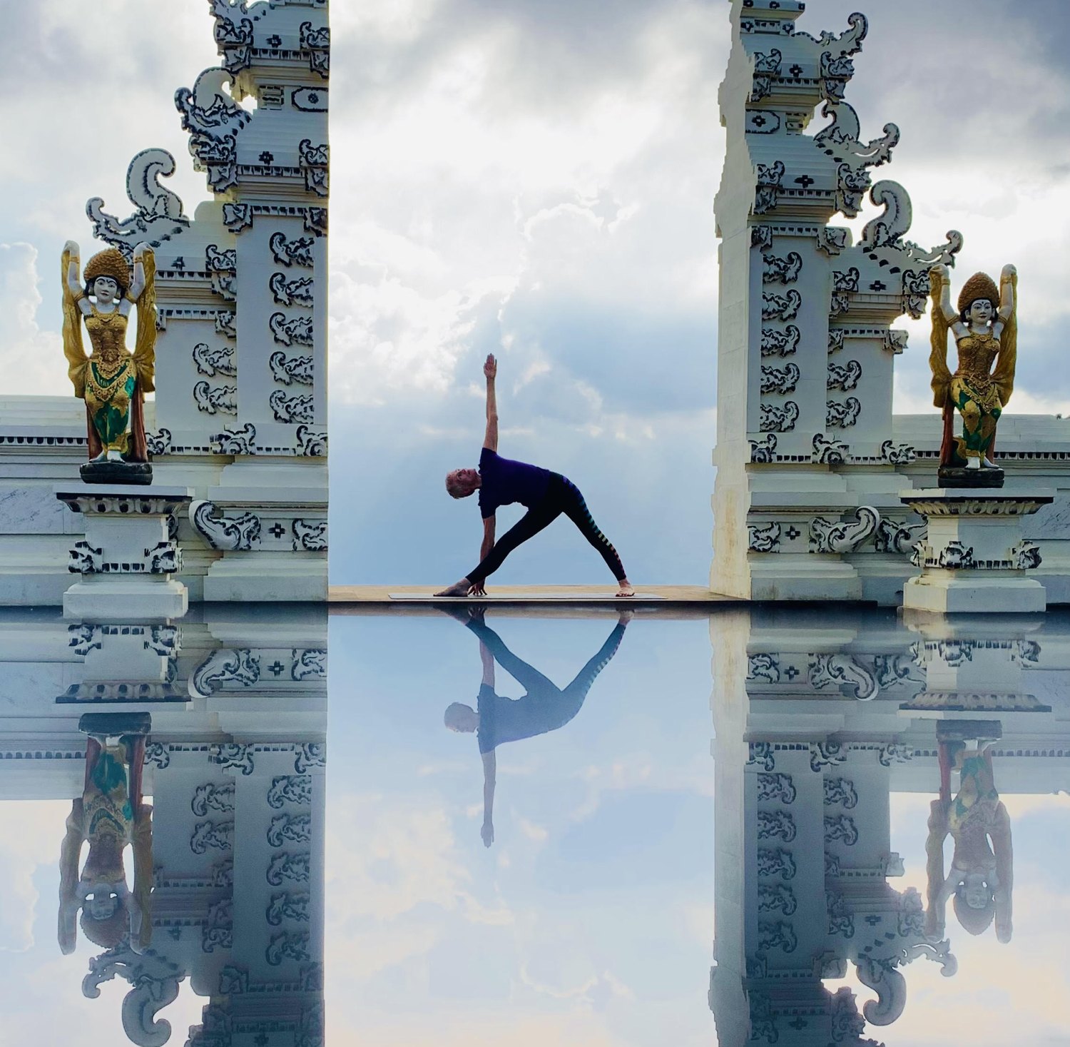 A Flametree teacher in Utthita Trikonasana on a mat between the carved white gates at the Lahangan Sweet lookout in east Bali, with the mirrored reflection below