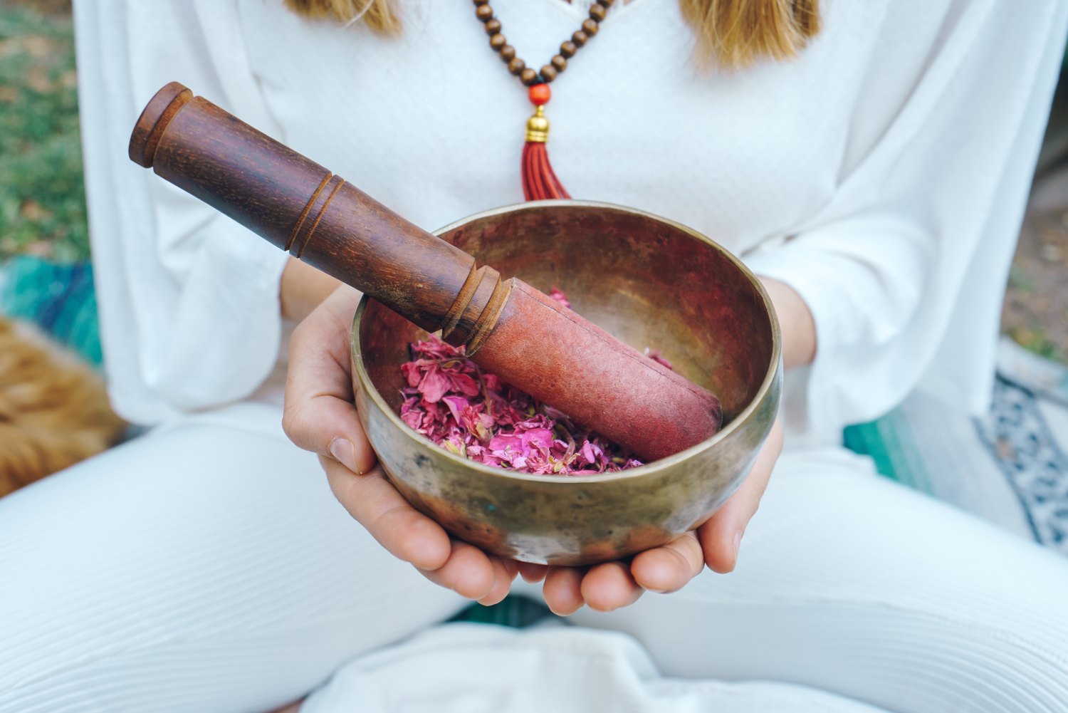 A woman holding a hand beaten Tibetan singing bowl with a wooden mallet and rose petals inside, sitting outdoors with greenery behind