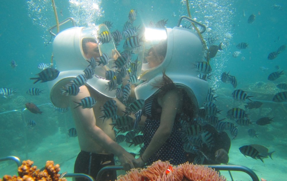 Two people wearing clear underwater helmets standing on the sea bed at Sanur, surrounded by sergeant major fish and coral