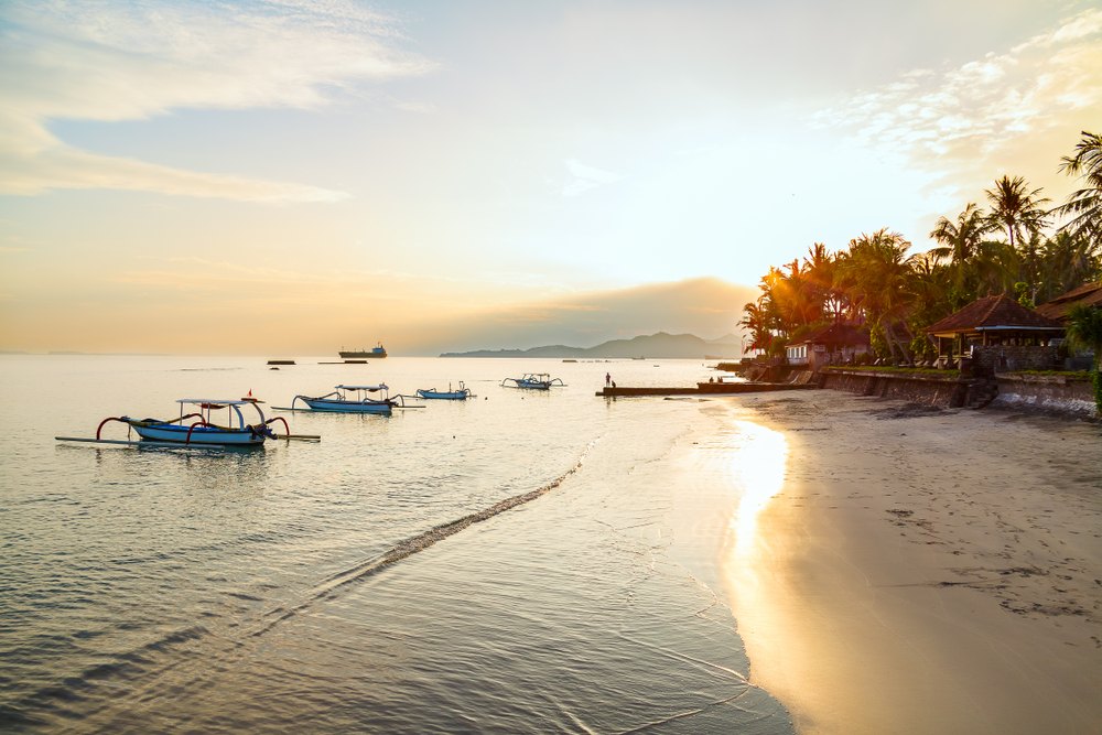 Several traditional jukung outrigger fishing boats moored in calm shallow water off the beach at Candidasa, with palms and low sun behind