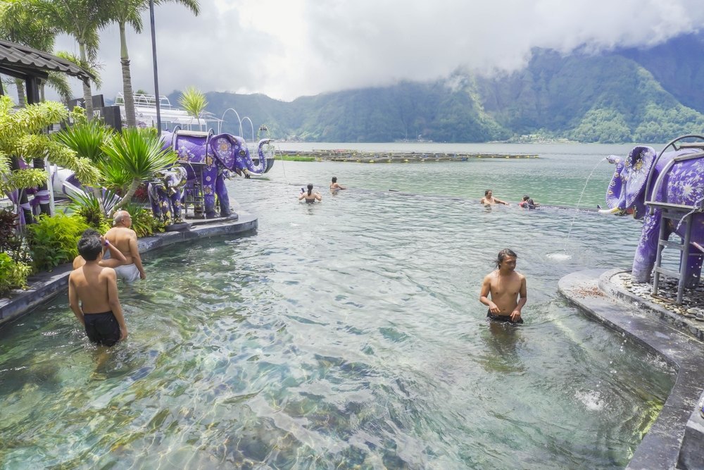 People sitting in the warm spring fed pools on the shore of Lake Batur, with the caldera wall and cloud behind