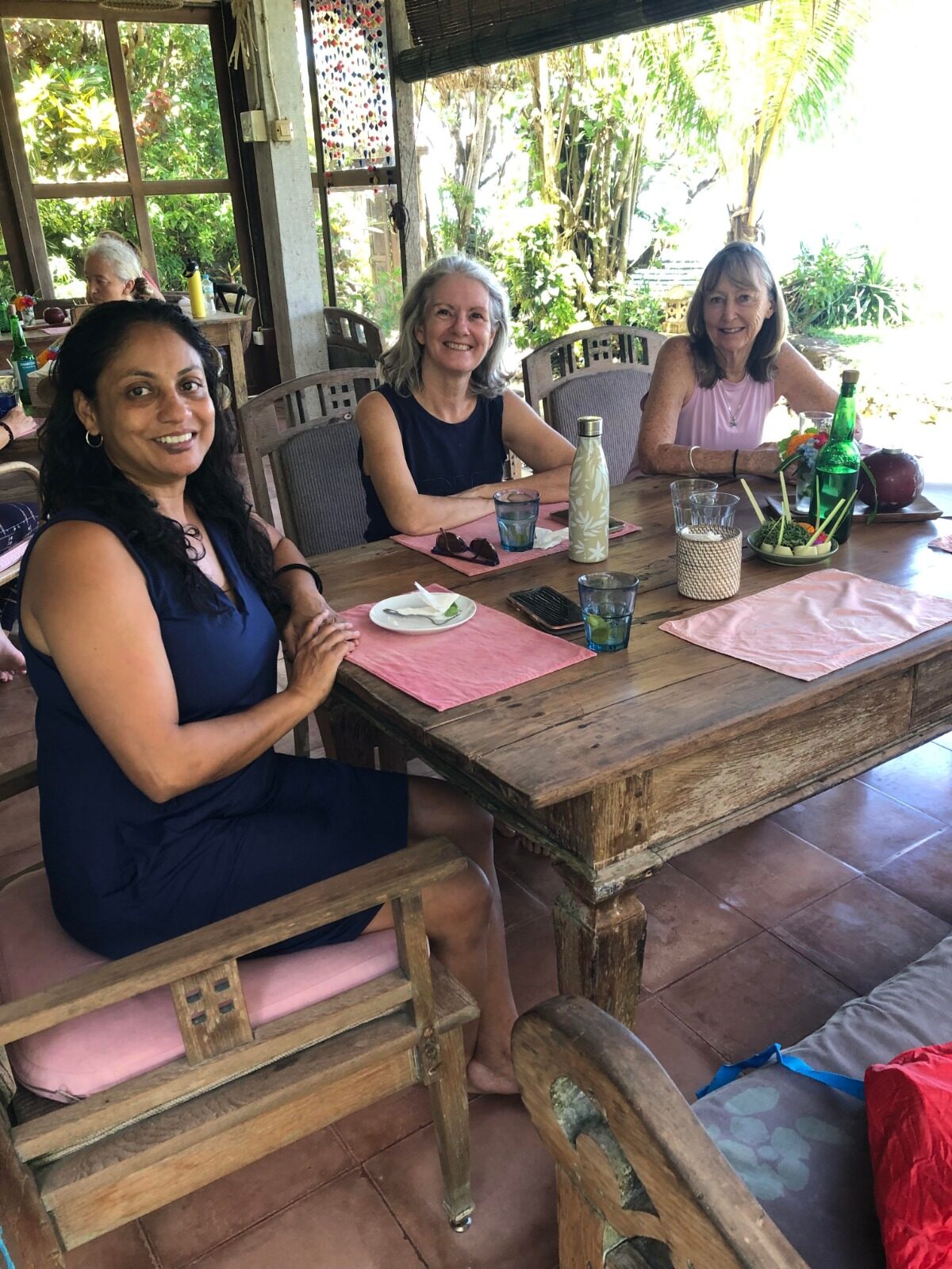Three women talking over lunch at a wooden table at a Flametree yoga retreat in Candidasa, Bali