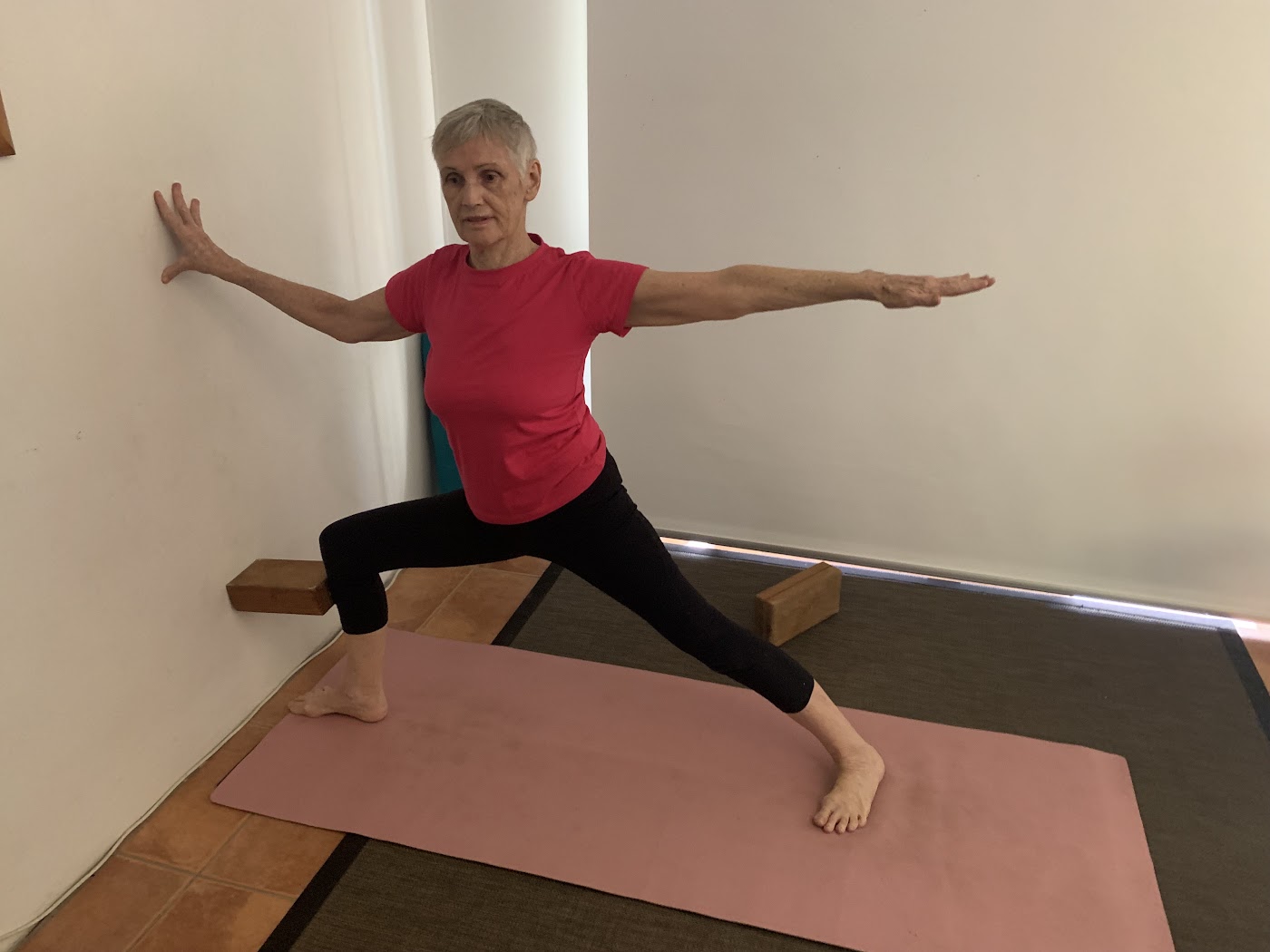 Older woman in Warrior 2 with her back hand on the wall and blocks beside her feet