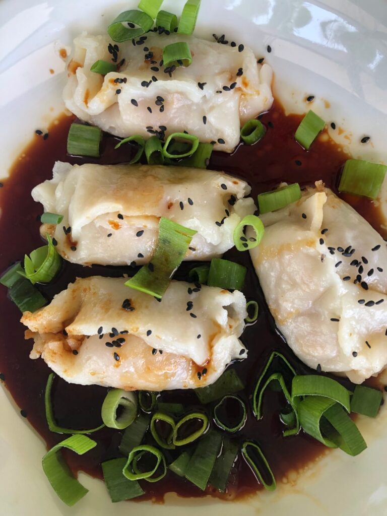 Steamed gluten free wontons on a white plate with dipping sauce, spring onions and sesame seeds