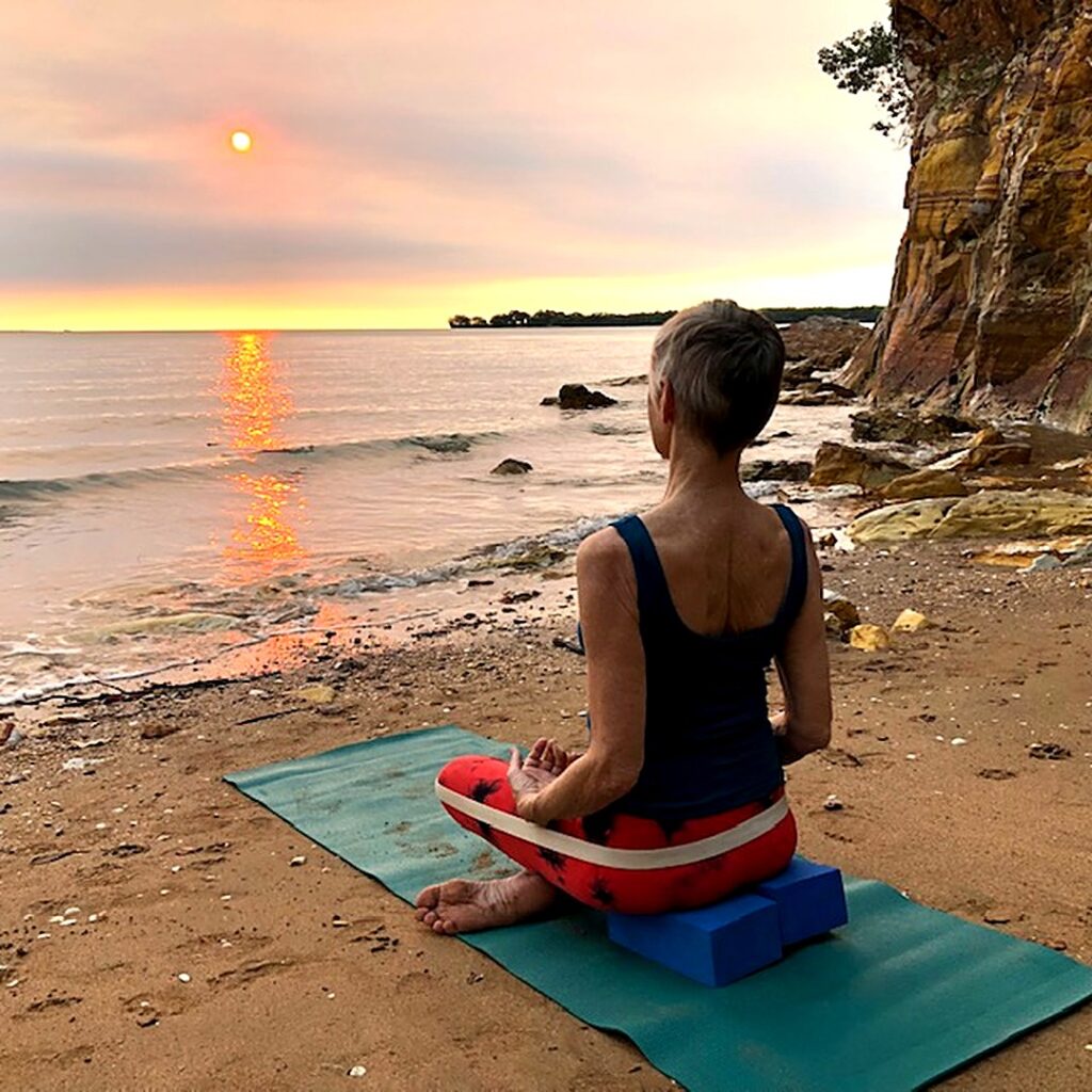 Piriformis stretch: Chris Lalor sitting tall on a block with a belt round her legs, weight on her sitting bones, by the sea in Darwin