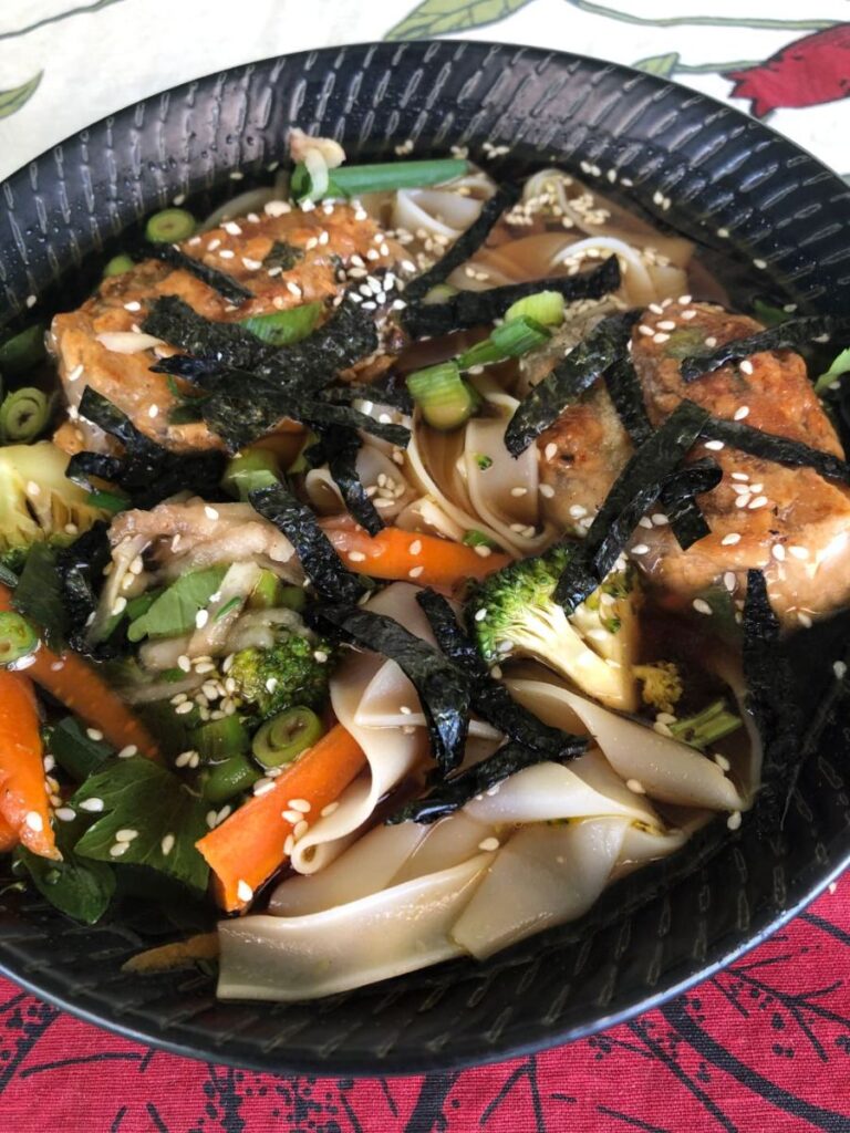 Rice noodle soup in a black bowl with tofu rice paper rolls, carrot, broccoli, nori strips and sesame seeds