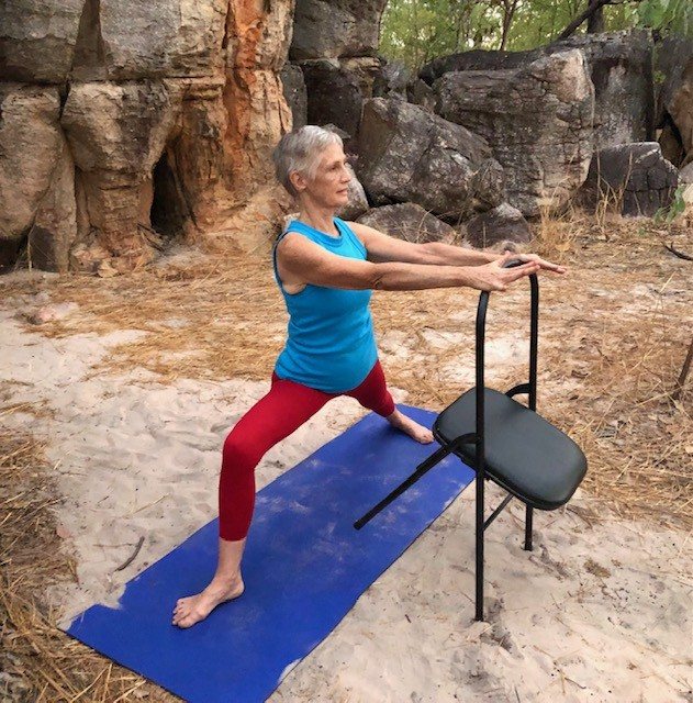 Osteopenia Darwin class: Chris Lalor in a standing lunge pose holding the back of a chair, loading the legs and hips on sand