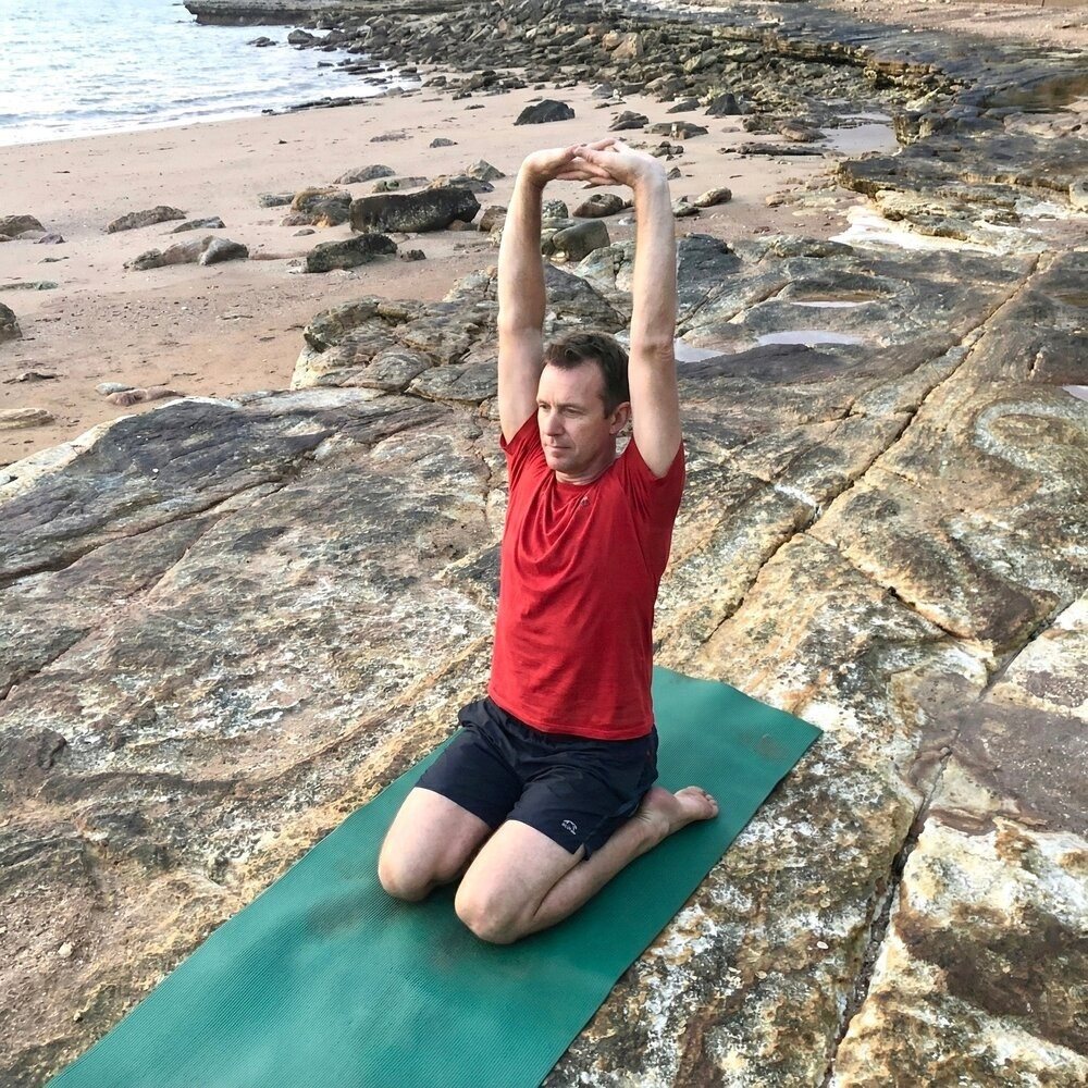 Stiff neck relief: a man kneeling on a mat with both arms stretched straight overhead, chest open, by the sea