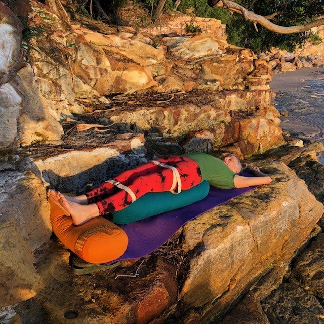 Yoga for perimenopause: Chris Lalor resting in a supported reclining pose on bolsters on a rock by water